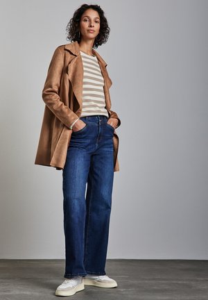 Beige Wildleder-Trenchcoat über einem gestreiften beige-weißen Hemd, kombiniert mit blauen weiten Jeans und weißen Sneakern. Einfarbiger Hintergrund.