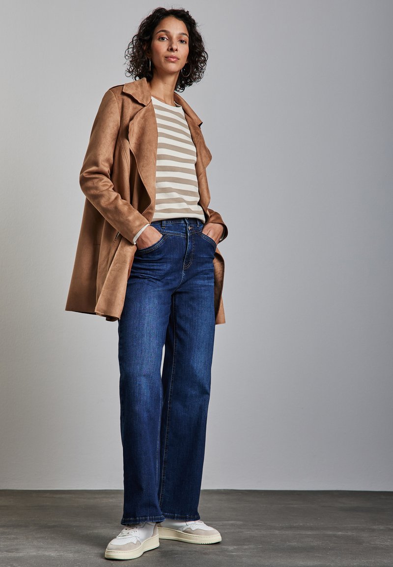 Trench coat de ante color marrón claro sobre una camisa de rayas beige y blanca, combinado con jeans de pernera ancha azul y zapatillas blancas. Fondo liso.