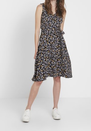 Donna che indossa un vestito nero senza maniche fino al ginocchio con stampa floreale bianca e gialla e sneakers bianche, in piedi davanti a uno sfondo semplice.