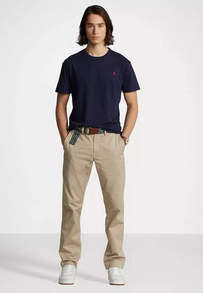 Maglietta a maniche corte blu navy con un piccolo logo, chinos beige e una cintura a righe. Scarpe bianche e un orologio completano l'outfit.