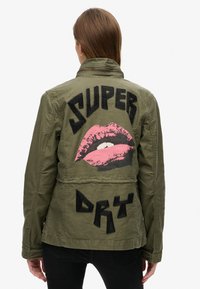 Olivengrøn jakke med et struktureret sort "SUPER" og "DRY" design, der har et stort pink læbe-grafik på bagsiden. Ensfarvet front, afslappet pasform.