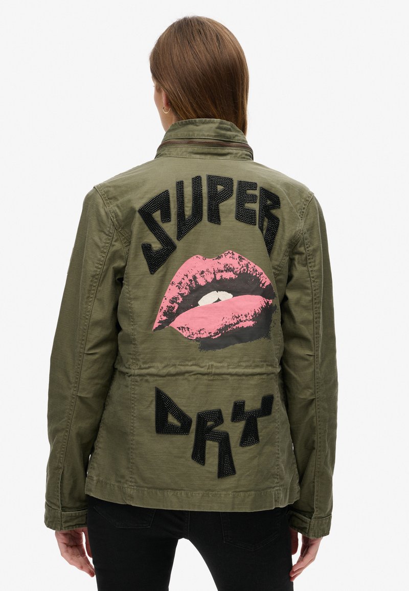 Olivengrøn jakke med et struktureret sort "SUPER" og "DRY" design, der har et stort pink læbe-grafik på bagsiden. Ensfarvet front, afslappet pasform.