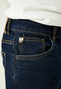 Jeans de mezclilla azul oscuro con costuras en contraste, un pequeño logo en el bolsillo frontal y un diseño clásico de cinco bolsillos.