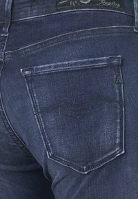 Jeans de color azul oscuro con un acabado de mezclilla texturizado, que presentan un bolsillo trasero con un diseño minimalista y un patrón de costura visible.