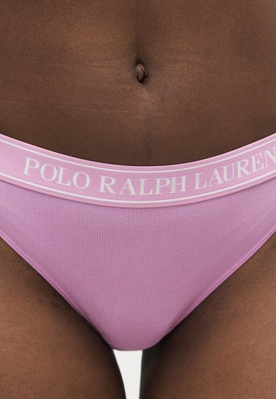 Άτομο με σκουρόχρωμο δέρμα φορεί ροζ ριμπ εσώρουχο με την επιγραφή «POLO RALPH LAUREN» σε λευκό στο λάστιχο, ορατός κορμός και άνω μηροί.