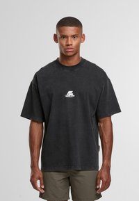 Mladý muž s krátkými vlasy má na sobě volné černé tričko s bílým logem a olivově zelené kraťasy, stojí na jednobarevném šedém pozadí.