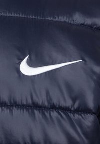 Μπλε επανωφόρι με γέμιση που διαθέτει το χαρακτηριστικό λευκό λογότυπο σφήνας της Nike. Απαλή, γυαλιστερή υφή με επενδεδυμένα τμήματα. Ελαφρύ σχέδιο.