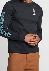 Hombre vestido con una camiseta de manga larga negra de Levi's con texto azul en las mangas y un gráfico pequeño cerca del pecho, acompañado de pantalones marrones.