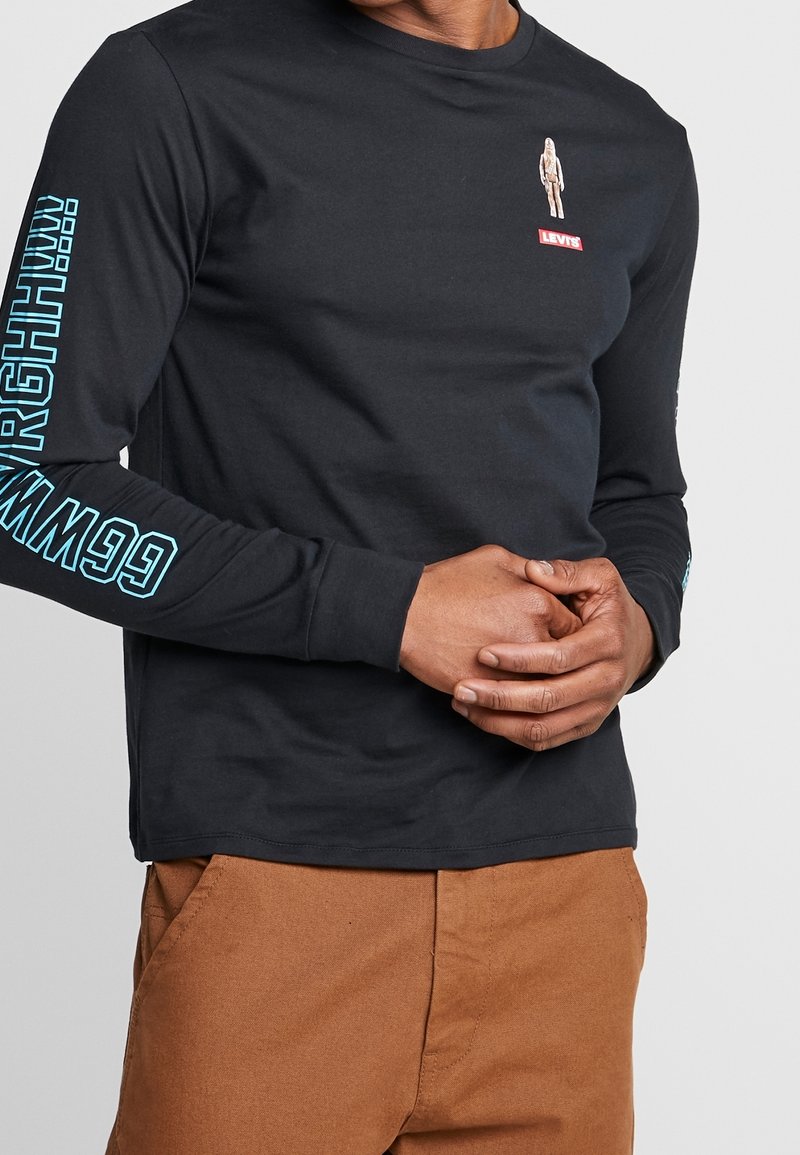 Hombre vestido con una camiseta de manga larga negra de Levi's con texto azul en las mangas y un gráfico pequeño cerca del pecho, acompañado de pantalones marrones.