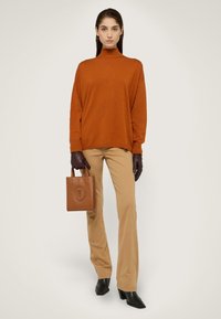 Ein rostfarbener Rollkragenpullover, beigefarbene Anzughose, braune Lederhandschuhe und eine braune Tote-Tasche mit geprägt designten Mustern. Schwarze Knöchelstiefel.