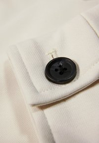 Krämfärgad tyg med en texturerad yta, med en svart fyrhålsknapp på en vikbar manschett. Sömnadsdetaljer är synliga nära knappen.