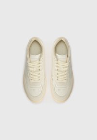 Cream-colored sneakers featuring a smooth upper material, rounded toe, textured accents, and flat laces. Rubber sole with a slight transparency.
