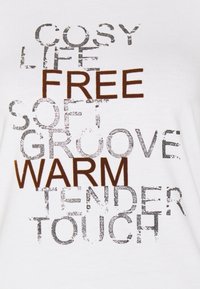 Weißes Baumwoll-T-Shirt mit variierenden Texten in Braun und metallic Silber, einschließlich Wörtern wie "KOSTENLOS", "WEICH" und "WARM", mit einem abgenutzten Look.