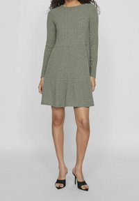 Femme portant une robe olive à manches longues, côtelée, atteignant le genou, avec des sandales à talons hauts noires à bout ouvert, sur un fond clair uni.