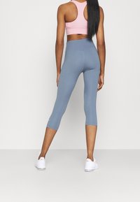 Valkoinen urheiluliivi ja siniset capri-lahkeet, istuva muotoilu, sileä tekstuuri, logotunnus. Yhdistetty valkoisiin urheilukenkiin.