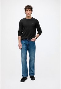 Homme debout avec une main dans la poche, portant une chemise noire à manches longues, un jean bleu, une ceinture noire, des chaussures noires et une montre-bracelet sur fond blanc.