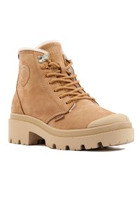 Bottines en suède beige avec bout arrondi, col rembourré et semelle robuste ; elles présentent des œillets en métal et un détail de logo sur le côté.