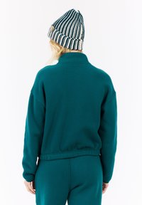 Persona di spalle che indossa una maglietta a maniche lunghe color turchese e pantaloni, con un berretto a maglia a strisce in bianco e turchese scuro.