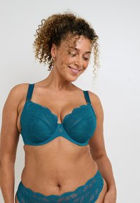 Reggiseno in pizzo teal con bordi scallop, coppe sagomate, spalline regolabili e dettaglio centrale rotondo in metallo. Tessuto morbido, elegante motivo floreale.