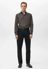 Camisa de botones de color marrón oscuro con un patrón texturizado, combinada con vaqueros negros de pierna recta y zapatos de vestir negros. Hombre de pie contra un fondo blanco.