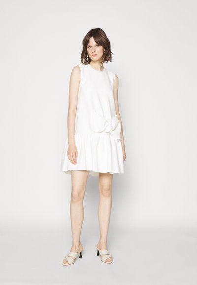Bijela haljina bez rukava s raskošnim rubom i velikom mašnom na struku. Mekani materijal s glatkom teksturom. Model nosi svijetle sandale.