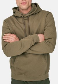 Oliwkowy hoodie z sznurkiem, kieszenią z przodu i ściągaczami na rękawach. Wykonany z miękkiego materiału o subtelnej teksturze i wykończeniach szytych.