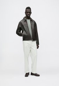 Vīrietis valkā tumši brūnu ādas jaku, gaiši pelēku džemperi, baltas bikses un tumši brūnas kurpes, stāvot pret vienkāršu gaišu fonu.