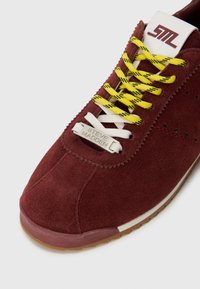 Sneaker in suede color bordeaux con lacci gialli e bianchi, etichetta in metallo "Steve Madden" per i lacci, e linguetta bianca con il logo stilizzato "SM".