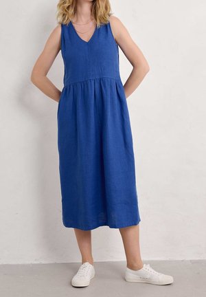 Femme portant une robe midi bleue sans manches avec un col en V et des baskets blanches, debout les mains derrière le dos devant un fond uni.