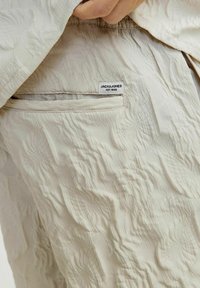 Pantalones de tela texturizada color beige con una superficie ondulada, que cuentan con un bolsillo trasero y una etiqueta que dice "JACK & JONES EST. 1990."
