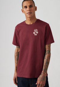 Bordeaux bomulds t-shirt med korte ærmer, rund hals, og broderet trælogo på brystet. Har en afslappet pasform og en glat tekstur.