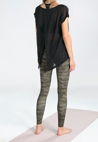 Top negro, de corte suelto y mangas cortas, con una hendidura en la parte posterior, combinado con leggings de camuflaje verde. Los materiales parecen ligeros y elásticos.