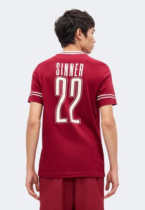 Jeune homme vu de dos portant un maillot de sport rouge avec des bandes blanches sur les manches et "SINNER 22" imprimé dans le dos.