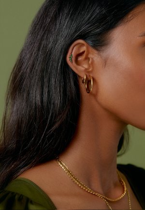 Pendientes de aro dorados y un pequeño pendiente de gema verde adornan la oreja, complementados por cadenas de oro en capas alrededor del cuello, sobre un fondo verde.