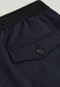 Marineblau gestreifter Stoff mit einem strukturierten Bund, der über eine kleine aufgenähte Tasche verfügt, die mit einem schwarzen Knopf gesichert ist. Dezenter Glanz und leichtes Material.