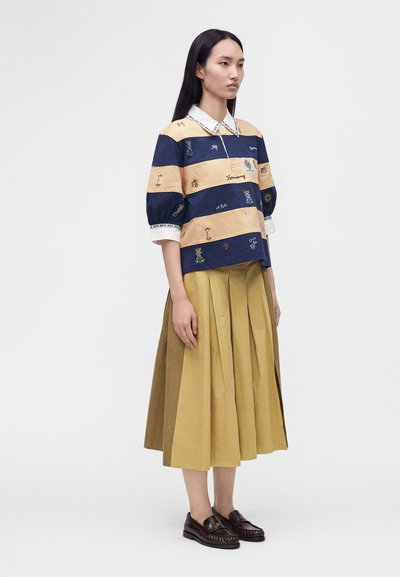 Femme aux longs cheveux foncés portant un haut brodé rayé bleu et beige, une jupe plissée jaune moutarde et des mocassins marron foncé, debout de profil.