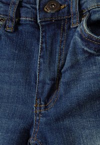 Tumši zila denim auduma, ar redzamām šuvēm, pogu aizdari un kabatām. Tekstūra ir nedaudz nodilusi, bet ar gludu virsmu.