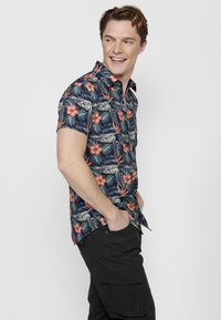 Korte mouwen marineblauw shirt met een bloemenpatroon in oranje en crème, met groene bladeren; gecombineerd met donkere cargo pants.
