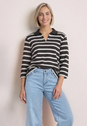Kvinde med kort blondt hår iført en sort og hvid stribet langærmet shirt og lyseblå jeans, smilende mod en lys baggrund.