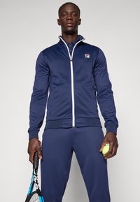 Mees tennisemängija tumesinises spordiriietuses, hoides ühes käes reketit ja teises tennispalli, seisab ühevärvilise tausta ees.