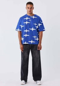 Camisa azul extragrande con patrones blancos en forma de estrellas, combinada con jeans oscuros de pierna ancha y zapatillas blancas. Diseño sencillo, ajuste relajado.