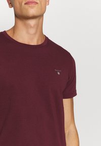 Man in een kobaltblauwe GANT T-shirt met korte mouwen en een klein logo op de linkerborst tegen een effen achtergrond.