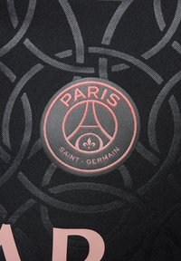 Logo del Paris Saint-Germain en rosa sobre un fondo de tela negra con un patrón circular y letras parcialmente en rosa claro debajo.