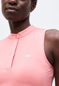 Κοντινό πλάνο ενός ατόμου που φοράει ροζ αμάνικο τοπ με μικρό λευκό λογότυπο κοντά στο στήθος και γιακά με κουμπιά.