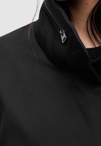 Zwarte jas met een hoge kraag en een zijrits. De stof heeft een subtiel geribd patroon en een gladde voering is zichtbaar bij de hals.