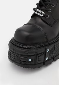 Svart läderboot med rund tå, tjock strukturerad sula och dekorativt hårdvara. Har metallsnörning och synliga sömmar.