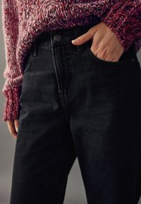 Schwarze hochtaillierte Jeans mit glatter Textur, ausgestattet mit einer Vordertasche und einem Knopfverschluss, kombiniert mit einem mehrfarbigen Strickpullover.