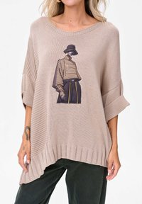 Jasno beżowy sweter z dzianiny w oversize'owym kroju, z nadrukiem przedstawiającym modela w paski oraz ściągaczami u dołu i na rękawach.