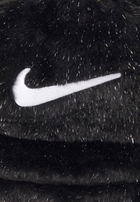 Melna vilnas cepure ar strukturētu virsmu, kurā ir spīdīgas šķiedras. Baltais Nike logo izcili redzams priekšpusē. Apaļa forma.
