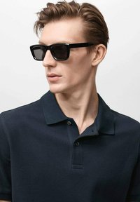 Námořnická polo košile se texturovaným materiálem, s knoflíkovou légou na tři knoflíky, nošena s černými obdélníkovými slunečními brýlemi. Hladký účes, neutrální pozadí.